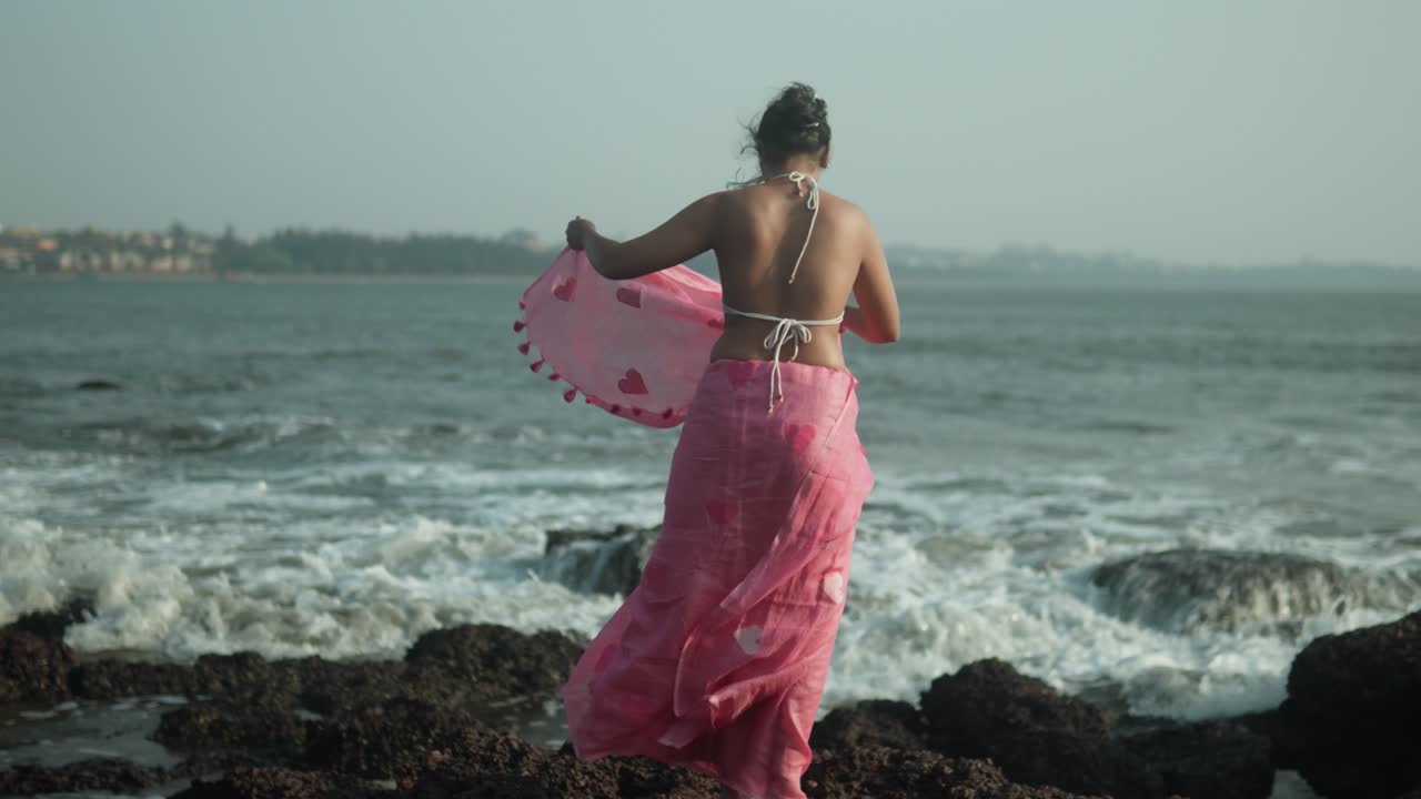 mujer con vestido rosa disfrutando de la brisa del mar en la orilla rocosa, las olas chocando detrás