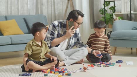 full body of asian father and sons playing the construction set colorful plastic toy brick at home. the father talks on smartphone watching and helping the kids assembling plastic building blocks