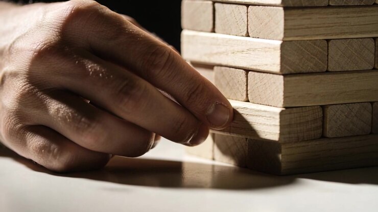 Mano che si allunga verso una torre, facendo scivolare un blocco di legno centrale su un tavolo con ombre intense.