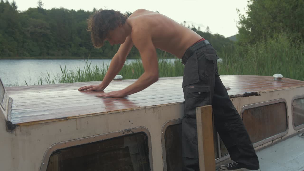 topless joven carpintero lijado a mano tablones de techo de barco de madera con papel de lija