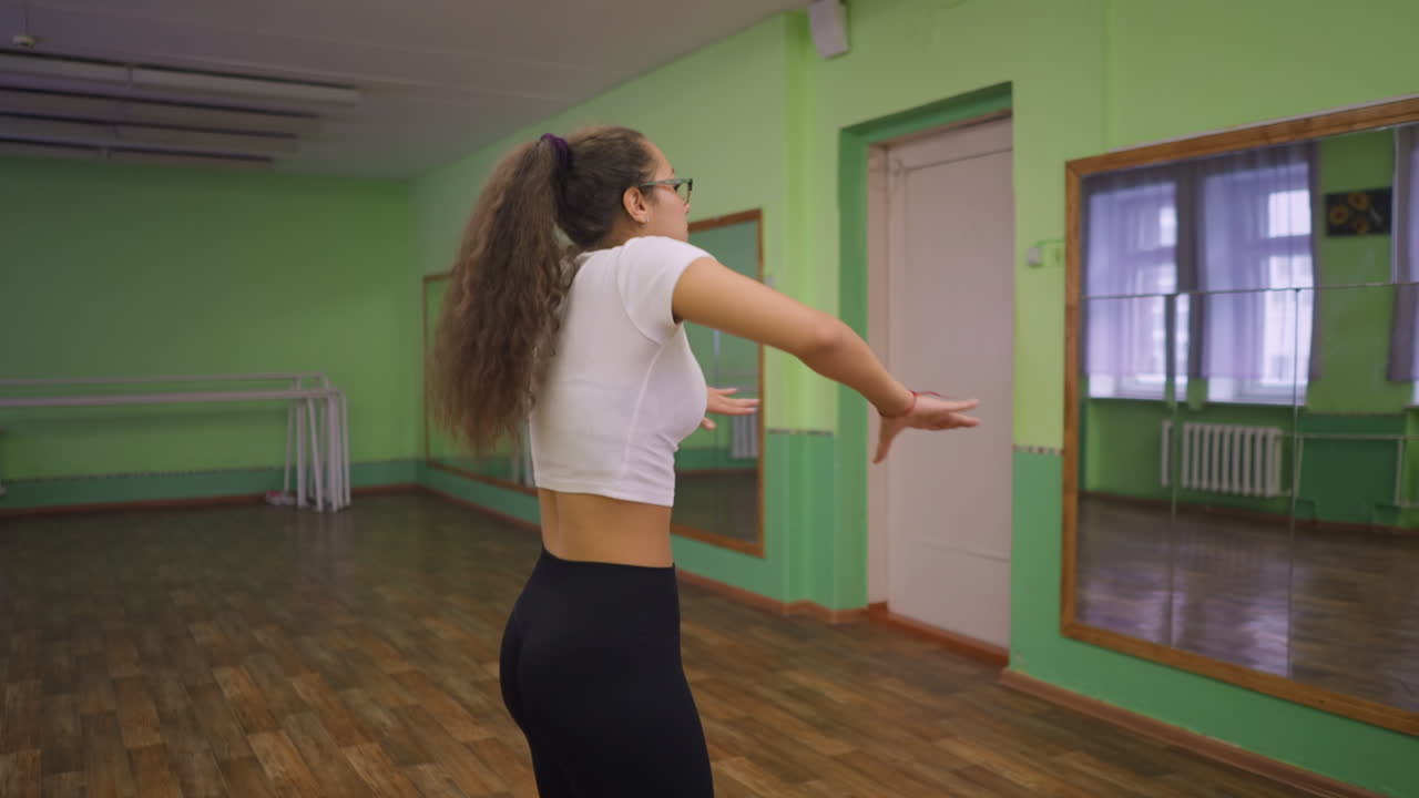 Close side view of woman in white top flipping her hair inside green painted room, background showing mirror reflecting her movements, wooden floor, and bright lighting creating energetic workout