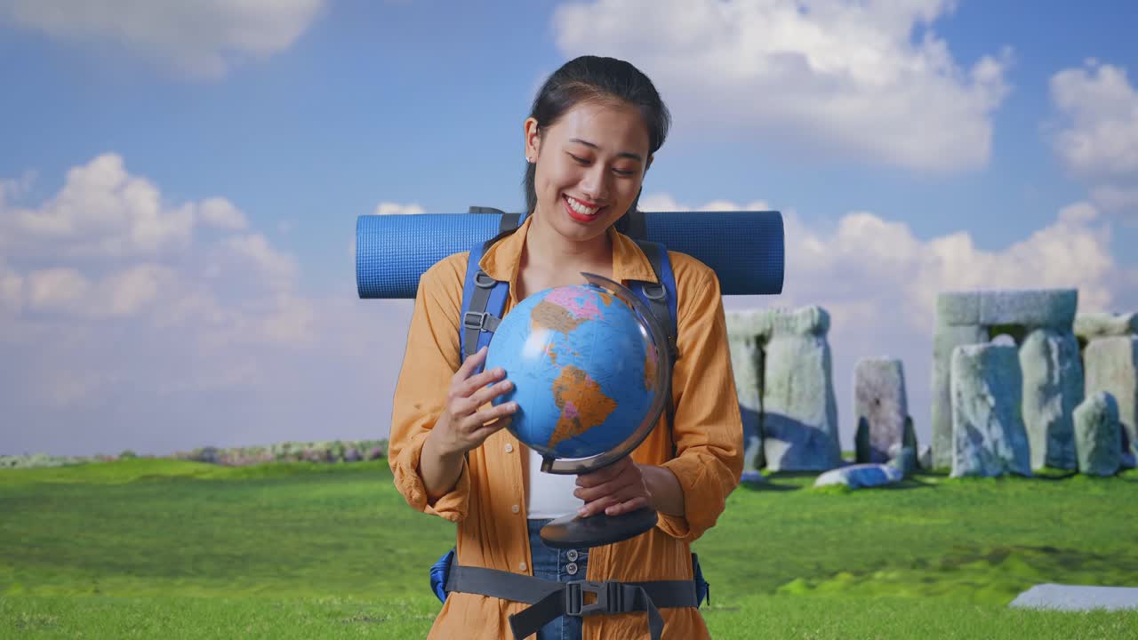 Asian Female Hiker With Mountaineering Backpack Holding World Globe In Her Hands And Smiling To Camera While Traveling In Stonehenge
