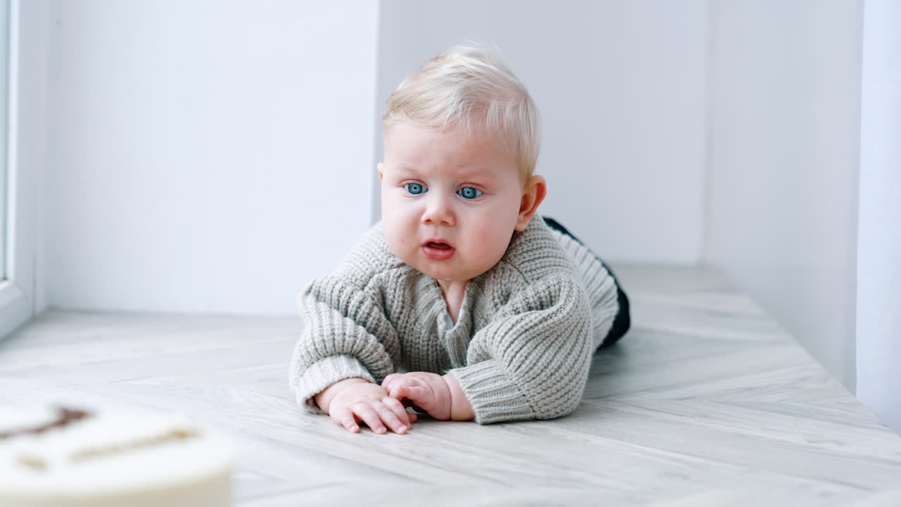 Lovely blond baby in grey sweater lies on his belly. Infant tries to crawl to the cake standing in front of him.