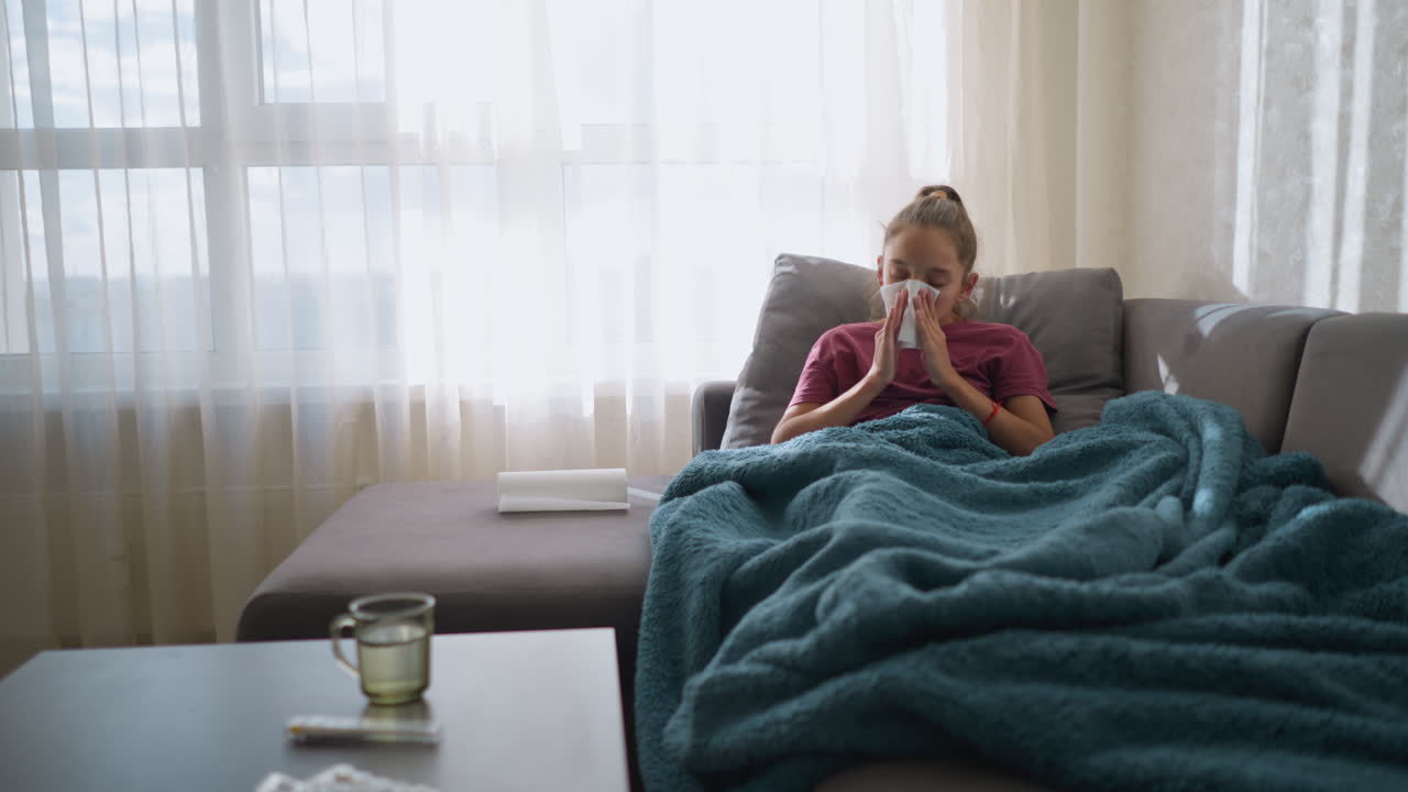 Sick girl sneezing while moving forward and back, tissue in hand, with medication on table, unwell, tired child covered in blanket, showing flu symptoms at home