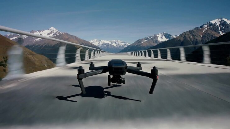 Rotors spinning hovering then moving black drone along viaduct toward snow peaks, gimbal visible