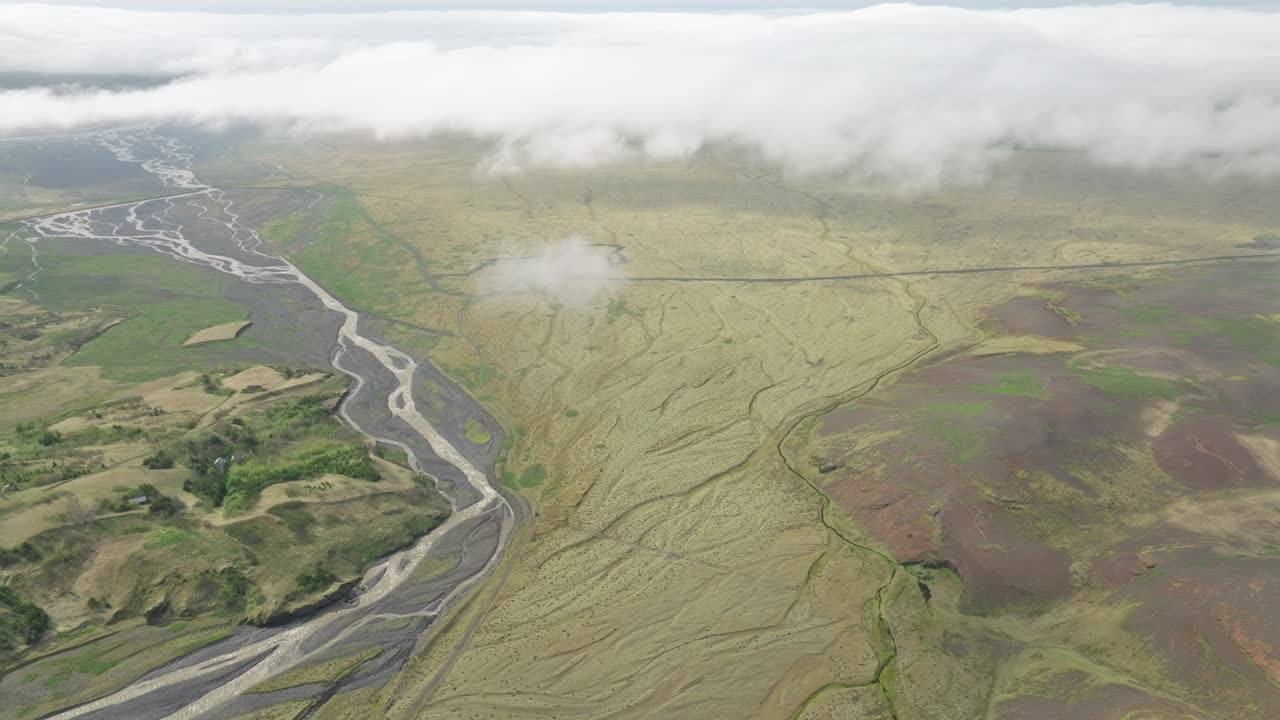 Aerial view of a braided river system flowing through expansive plains in Hafursey, Iceland, under low clouds, with patches of green terrain and volcanic features.