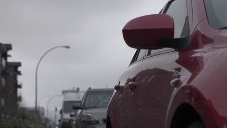 right side of a japanese red hatchback parked on the street in residential area on a cloudy fall day with traffic passing by in the background