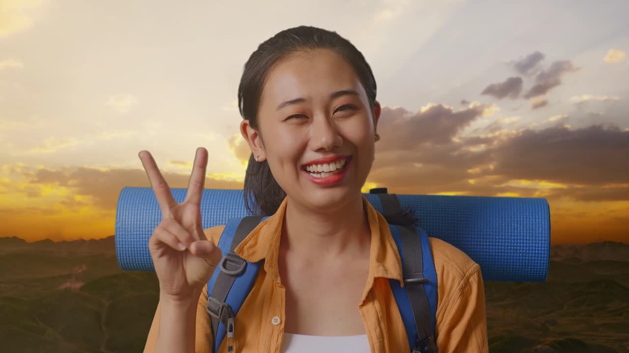 Close Up Of Asian Female Hiker With Mountaineering Backpack Smiling And Showing Peace Gesture While Standing On The Top Of Mountain During Sunset Time