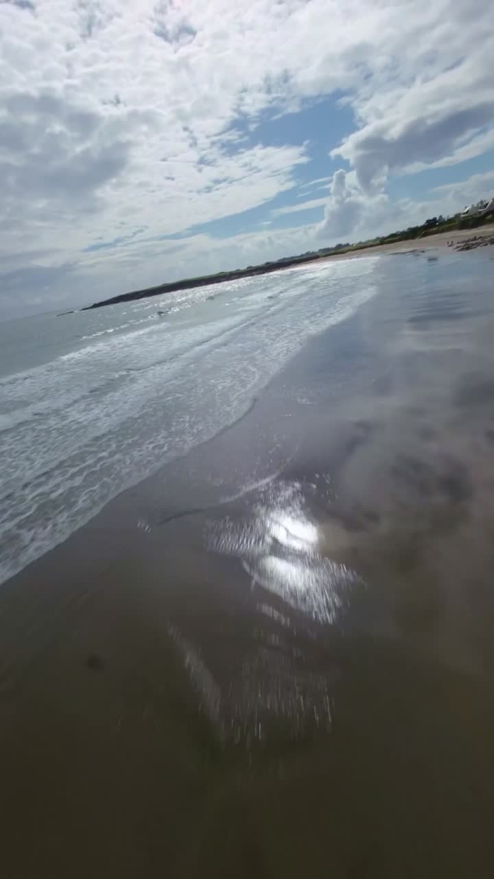 FPV drone descends along France Atlantic coast, Brittany cliffs drop to golden beaches where turquoise waves meet green slopes under partly cloudy sky