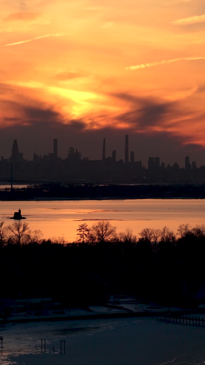 Dramatic golden sky glowing at sunset over the silhouette of the new york city skyline, with the sun's reflection shimmering on the water and a frozen shoreline in the foreground during winter