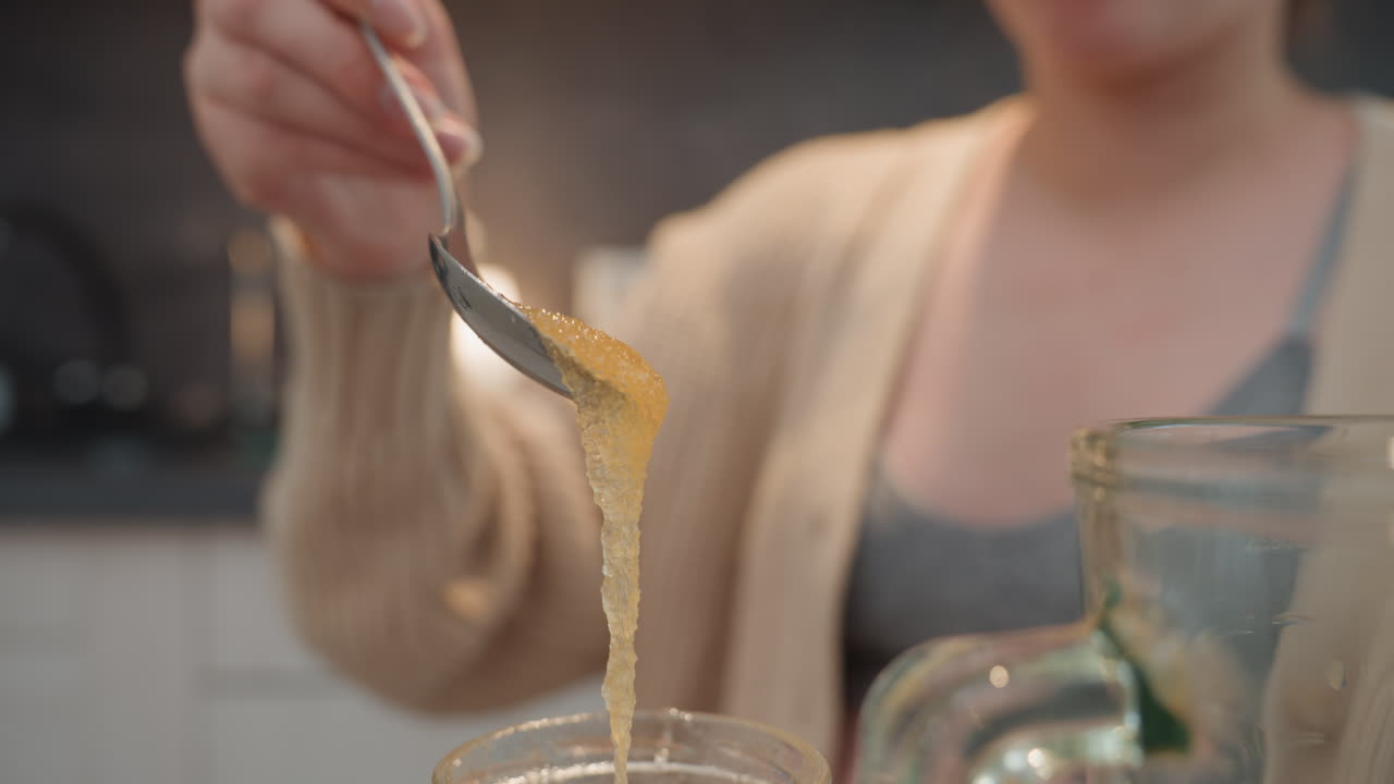 Donna incinta che mescola il miele in cucina, luce soffusa del mattino, cardigan accogliente, barattolo di vetro e frullatore, lento versamento di miele dorato in una ciotola, primo piano di mani e busto, routine di cura prenatale, nutriente