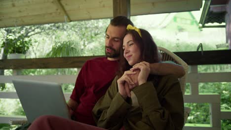 el chico y la chica se sientan y se balancean en una mecedora, la chica sostiene la mano del chico en sus manos. se comunican y ven algo en la pantalla de la computadora portátil en el gazebo con el telón de fondo de un bosque verde