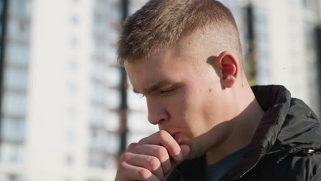close up of white boy bringing vape device to lips while inhaling with focused expression under bright daylight wearing black puffer vest and t-shirt with blurred urban background behind