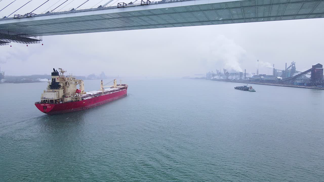 Drone follow of red cargo ship under bridge with Zug Island steel mill in background
