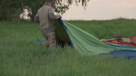 man standing in tall grass unfolding colorful hot air balloon envelope during preflight setup at sunset field, wearing cap, preparing fabric for inflation with teamwork vibe in rural landscape