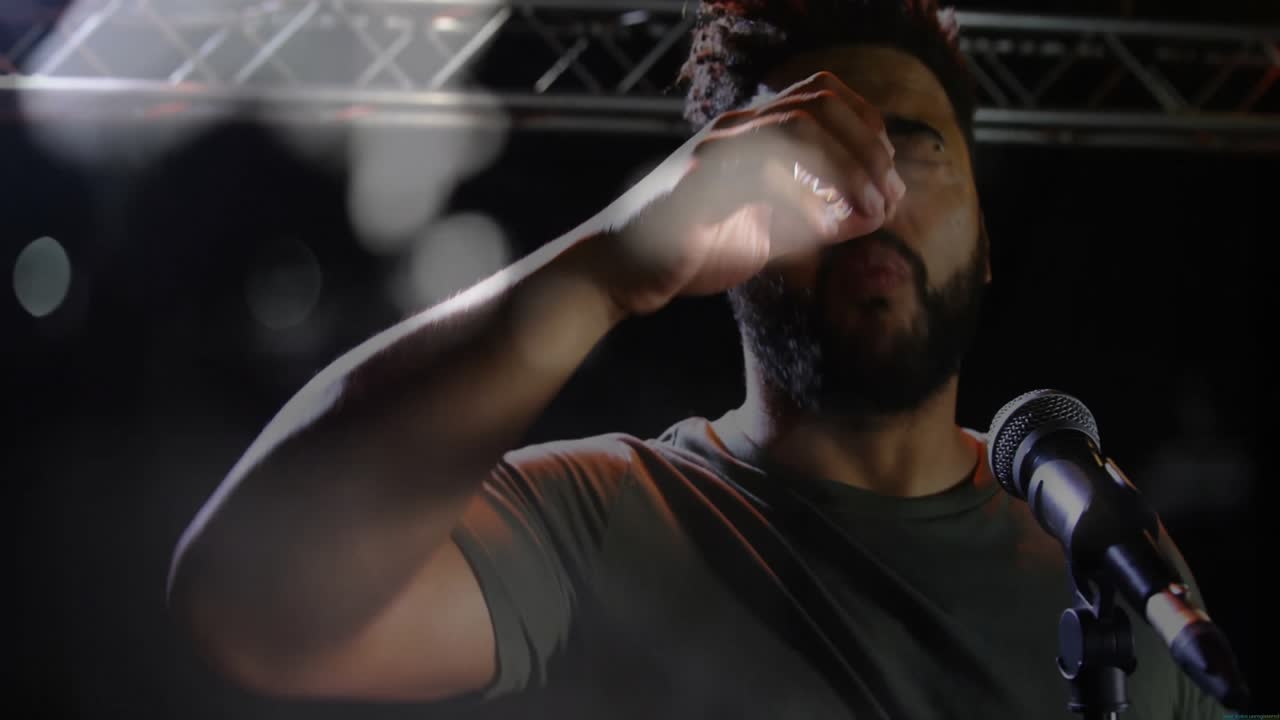 Male performer wiping sweat on music stage, with audio waves pulsing around mic under hazy lights