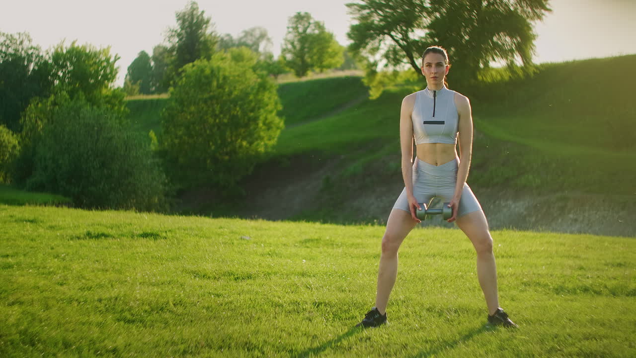 Women's training in the park. Girl squats with dumbbells in her hands at dawn standing on grass