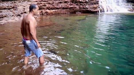 young man jumping and swimming in blue clear water of waterfall from flat angle