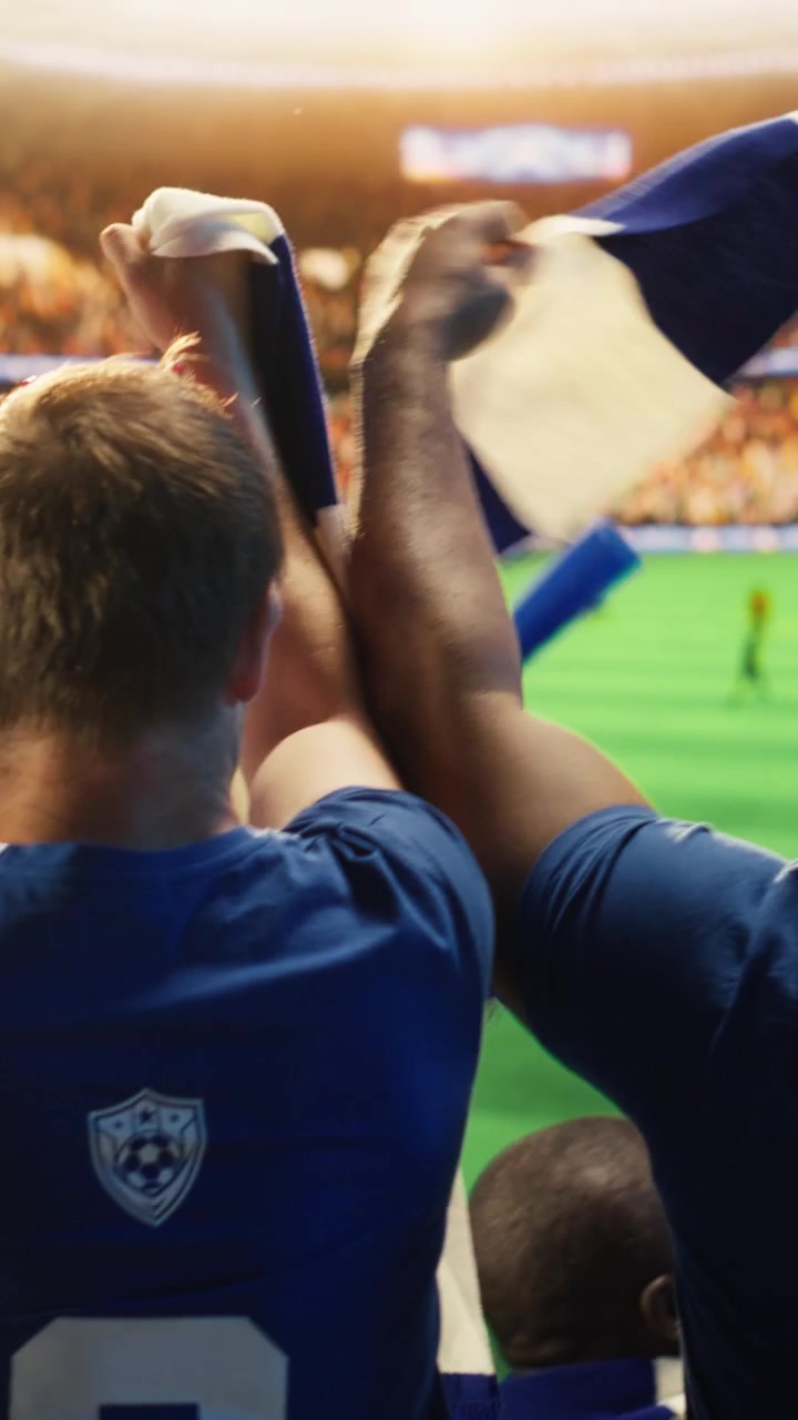Video vertical. Primer plano de hinchas de fútbol sosteniendo bufandas en alto durante un partido de fútbol.
