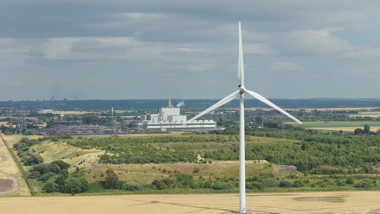 A tall wind turbine operates in a vast golden field under a cloudy sky. Its blades spin, harnessing wind energy. In the distance, an industrial complex is visible.