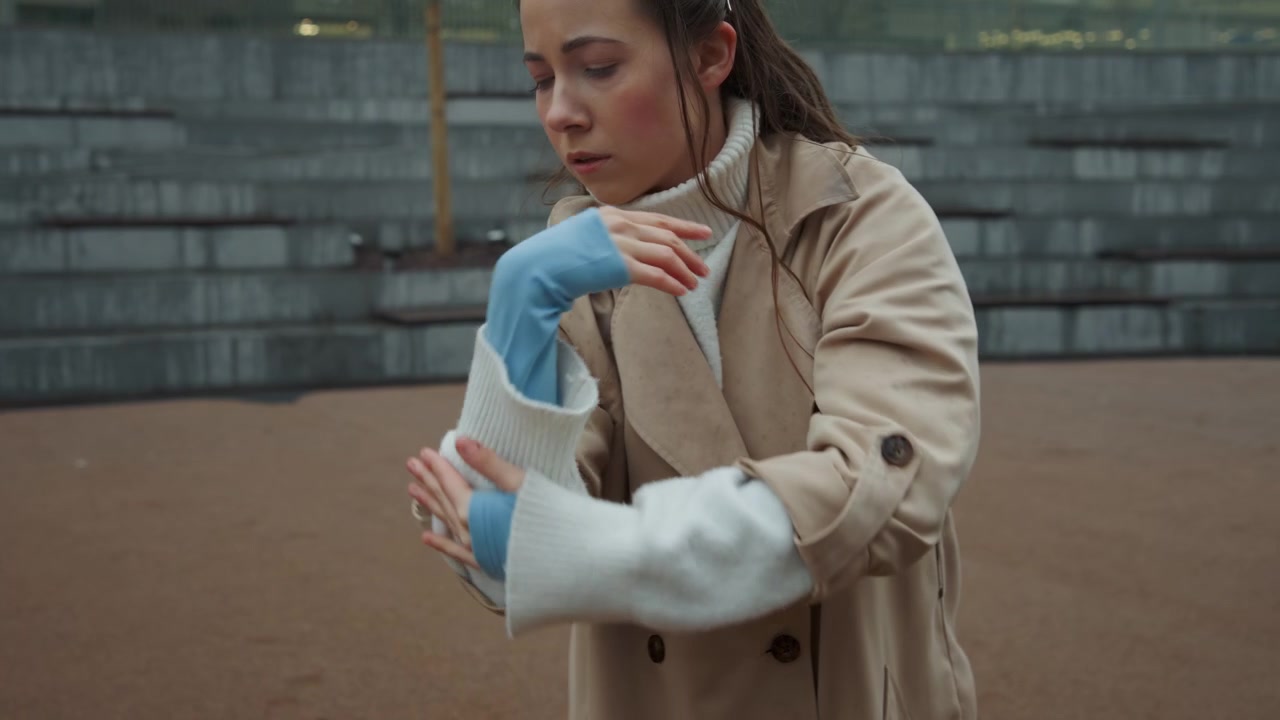 Primo piano di una giovane donna che sistema i suoi guanti bianchi a maglia e le maniche blu in un ambiente urbano esterno con scalini in pietra, edifici e alberi. La donna indossa un cappotto beige sopra abiti a strati, concentrandosi.