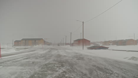 A-car-vanishes-into-the-distance-while-the-wind-moves-snow-accross-an-empty-road-in-Longyearbyen,-Svalbard