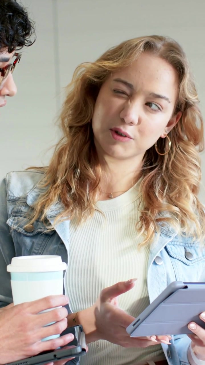 Video verticale: Uomo appoggiato che mostra un tablet, donna in jeans che discute con telefono e caffè in corridoio.