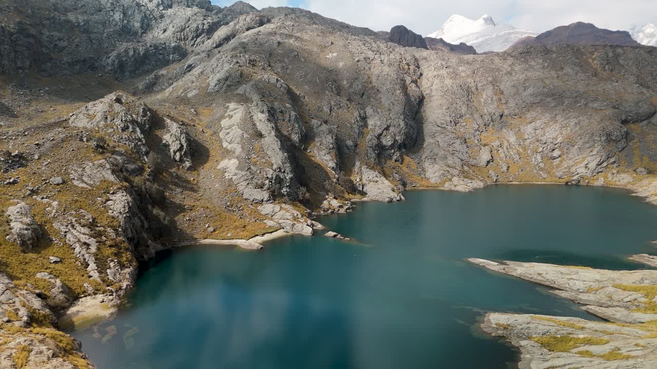 A stunning aerial flyover reveals a pristine, deep blue alpine lake at Paso del Zorro, nestled among the rugged, rocky peaks of the remote Huayhuash mountain range in the Peruvian Andes