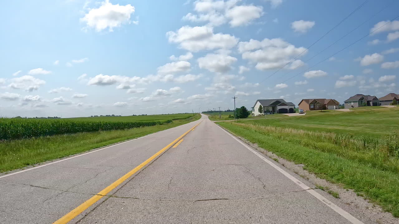 POV - driving on a country road past a few homes, power lines and fields full of crops in the field in late summer in the Des Moines River valley in central Iowa; concepts of farming and rural life