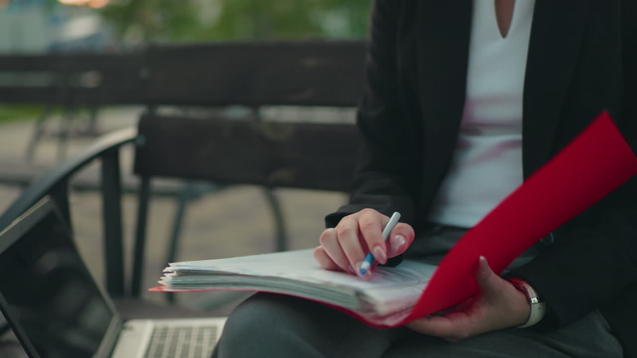 Close up of lady in professional attire flipping through red folder with pen in hand, seated next to laptop outdoors, with blurred bench and greenery in background