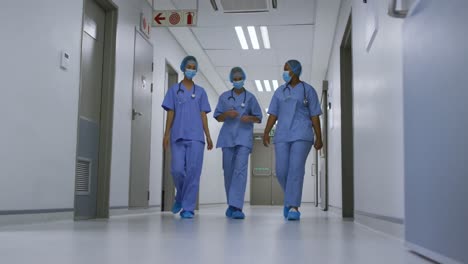 Diverse-female-doctors-wearing-face-masks-talking-walking-in-hospital-corridor