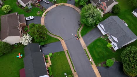cul-du-sac of american neighborhood with single family houses and waving green trees. rainy and cloudy day in spring. aerial top down shot. parking cars on driveway.