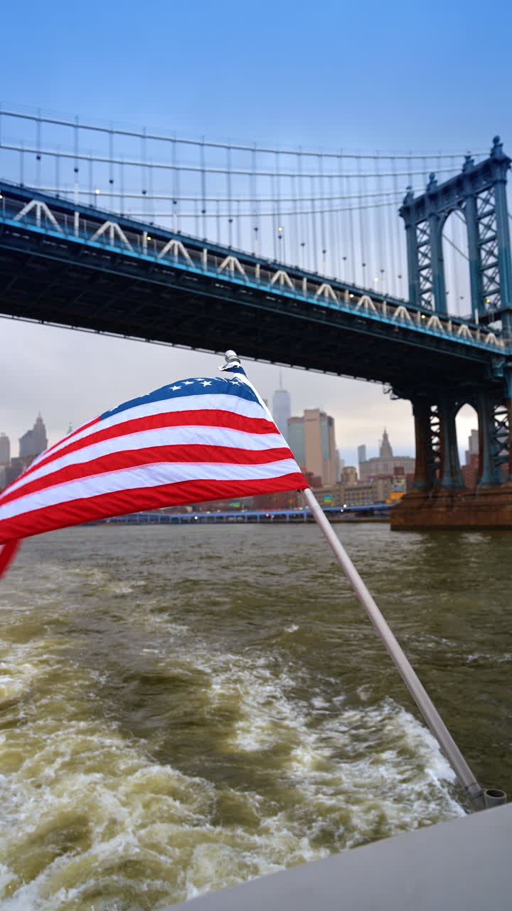 Bandiera americana che sventola su una barca vicino al Ponte di Manhattan, NYC. Bandiera degli Stati Uniti in primo piano con un ponte sospeso in acciaio blu e lo skyline della città sull'acqua.