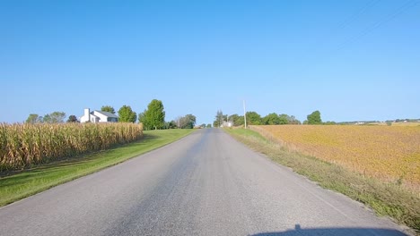 Pov-Conduciendo-En-La-Carretera-Rural-Del-Condado-Pasando-Por-Campos-Maduros,-Corrales-Y-Ciclistas-En-La-Zona-Rural-De-Iowa-En-Un-Soleado-Día-De-Otoño-Temprano