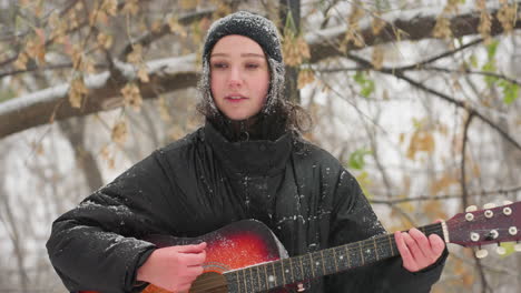 woman in black hoodie playing acoustic guitar outdoors in snowy park, surrounded by frosty bare trees and softly falling snow, capturing serene winter mood and harmony between music