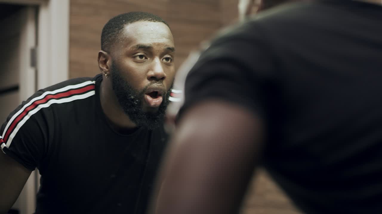 Black man washing face in luxury bathroom. Exhausted male looking at mirror.