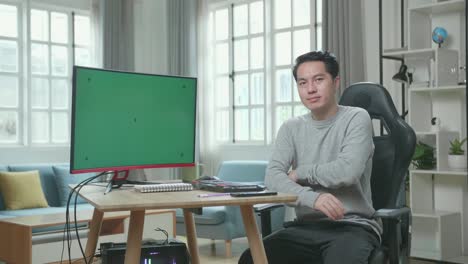 asian man in long sleeved t-shirt and black pants with green screen desktop computer crossing his arms and smiling to camera while working at home.