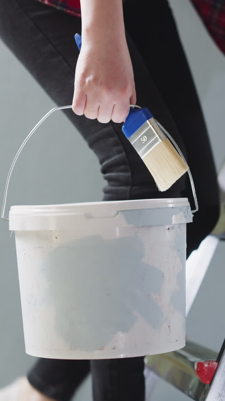 Woman goes up step ladder holding paint brush and bucket in room closeup. House owner does repair in apartment. Woman prepare to paint walls at home
