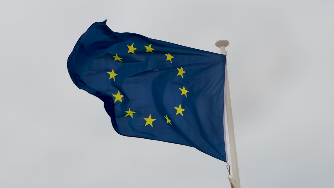 La bandiera dell'Unione Europea sventola energicamente su un pennone con un cielo nuvoloso sullo sfondo, telecamera statica, luce naturale