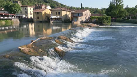 water flows over smooth wide shallow waterfall in borghetto verona italy, aerial dolly