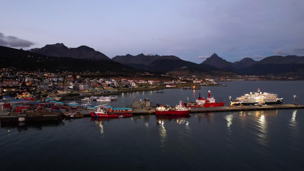 Aerial Drone Footage Captures The Stunning Ushuaia Village In Argentina During Summer Sunset. The Camera Pans Over The Bustling Port, Showcasing Ice-Breaking Ships, A Cruise Ship, And Mountains in 4K