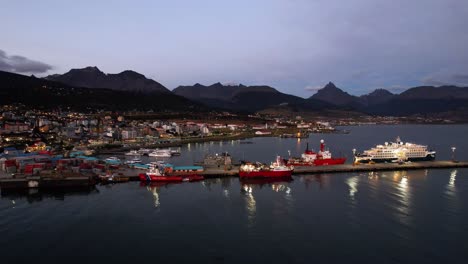 aerial drone footage captures the stunning ushuaia village in argentina during summer sunset. the camera pans over the bustling port, showcasing ice-breaking ships, a cruise ship, and mountains in 4k