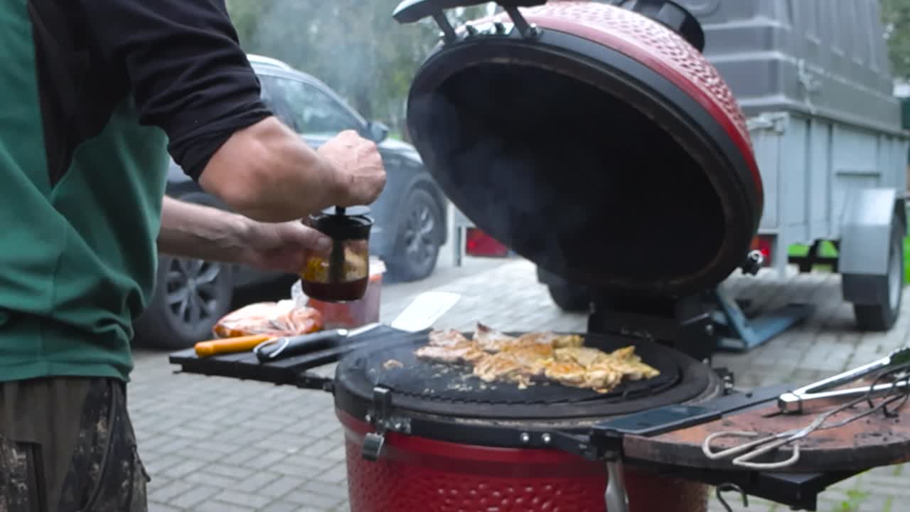 Uomo che spalma la salsa su carne o pollo alla griglia su una griglia a uovo rossa Komono o barbecue al rallentatore durante il giorno in estate o autunno. Mano maschile caucasica che condisce e insaporisce la carne su una griglia fumante