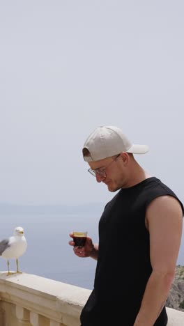 vertical shot of male drink espresso near seaside balcony with seagull aside