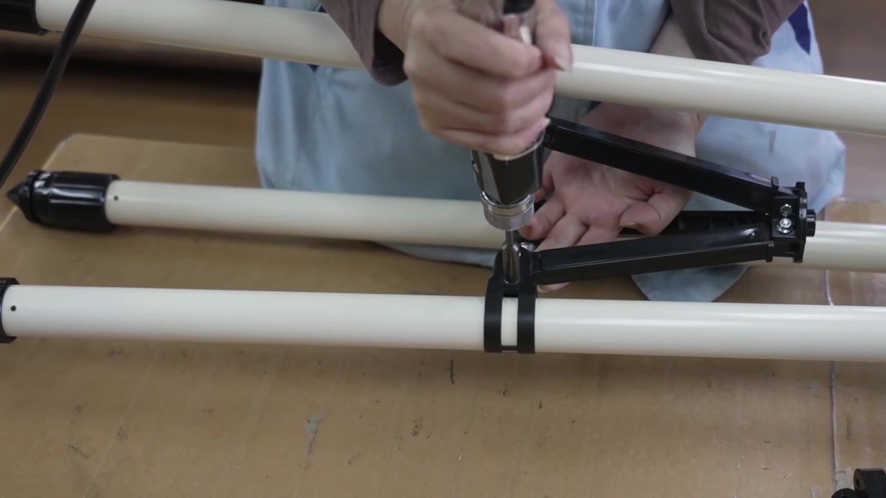 Close-up of worker attaching black tube ring clamp bracket to white telescope tripod leg using power drill