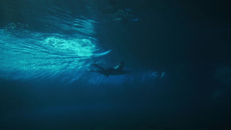 surfer dives below into deep water as barreling wave passes over with light rays shining through surface of ocean