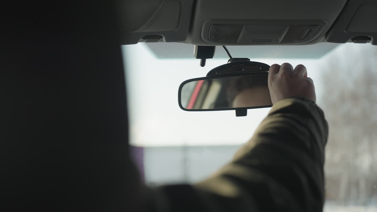 rear view of person in winter coat adjusting rearview mirror inside car, hand reaching upward with soft winter daylight streaming through windshield, exterior buildings visible in background