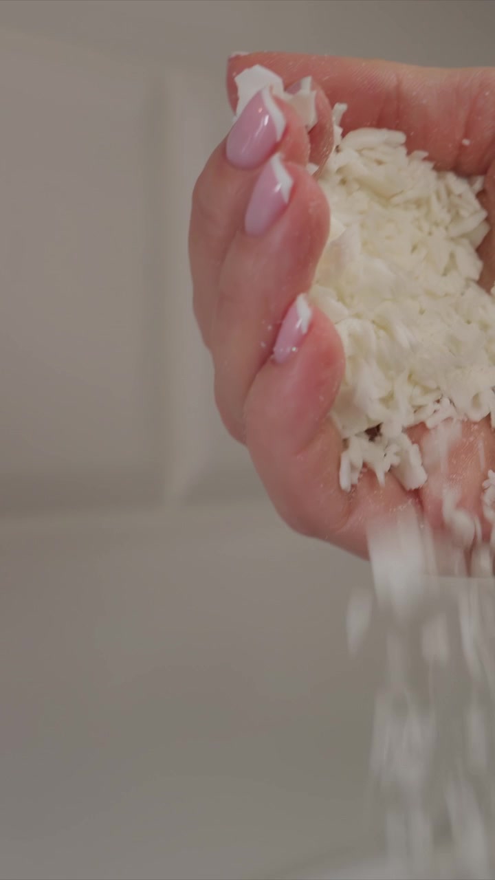 Primo piano di una mano con unghie rosa che tiene e rilascia materiale bianco sminuzzato sopra una ciotola con illuminazione soffusa. L'attenzione rimane sui dettagli della texture mentre piccole particelle si disperdono, enfatizzando l'interazione.