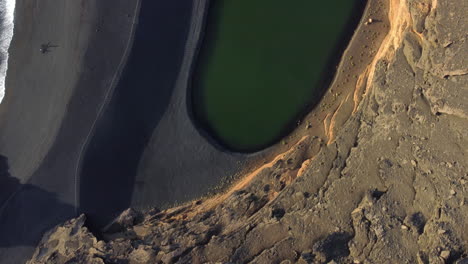 Beach-with-waves-and-rocks-next-to-a-lake-with-green-water,-black-sand-and-yellow-mountains