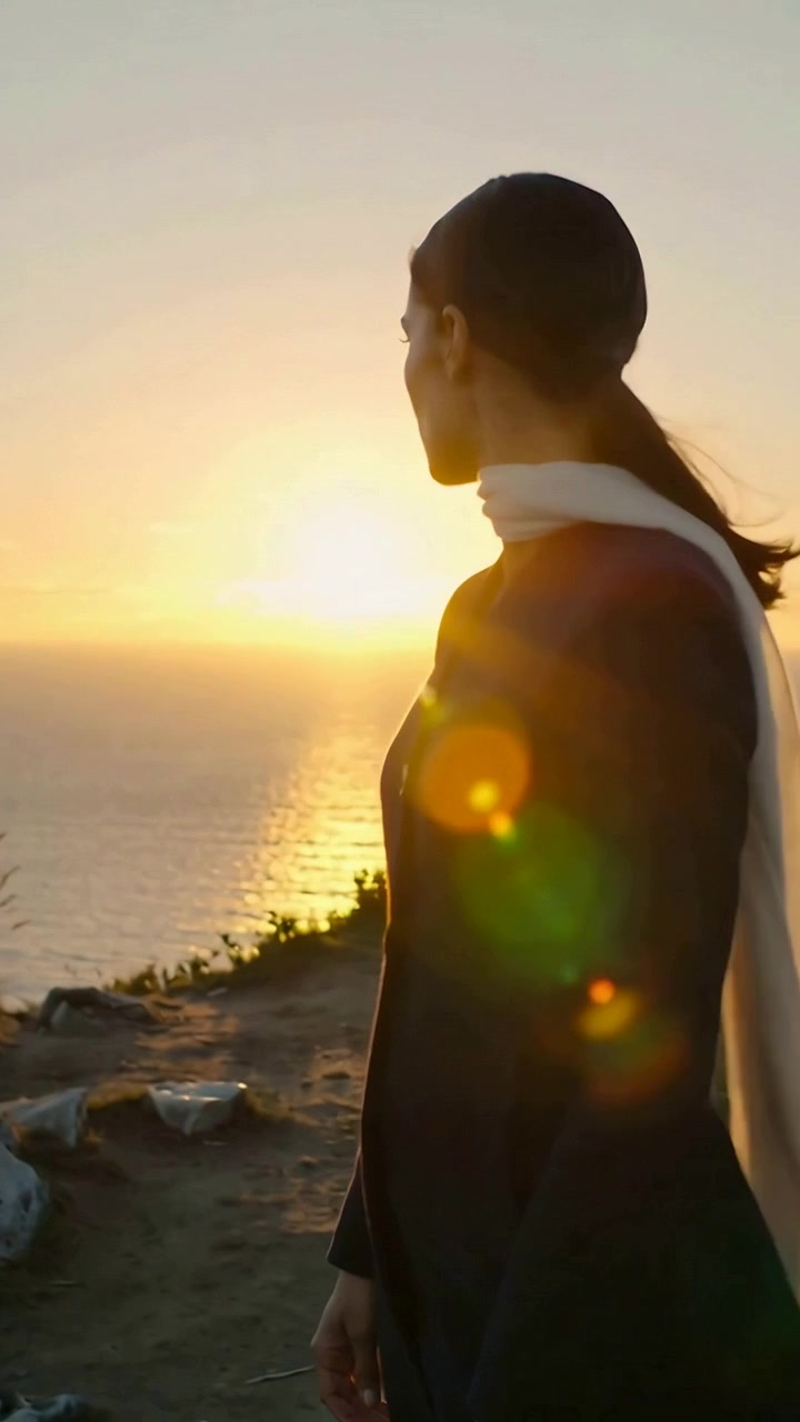 Video verticale: Donna con cappotto scuro che si gira verso il sole basso all'orizzonte su una scogliera, con la sciarpa che sventola.